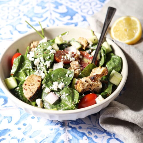 Side angle of a bowl of chicken shawarma salad on a blue and white tiled surface next to a neutral linen