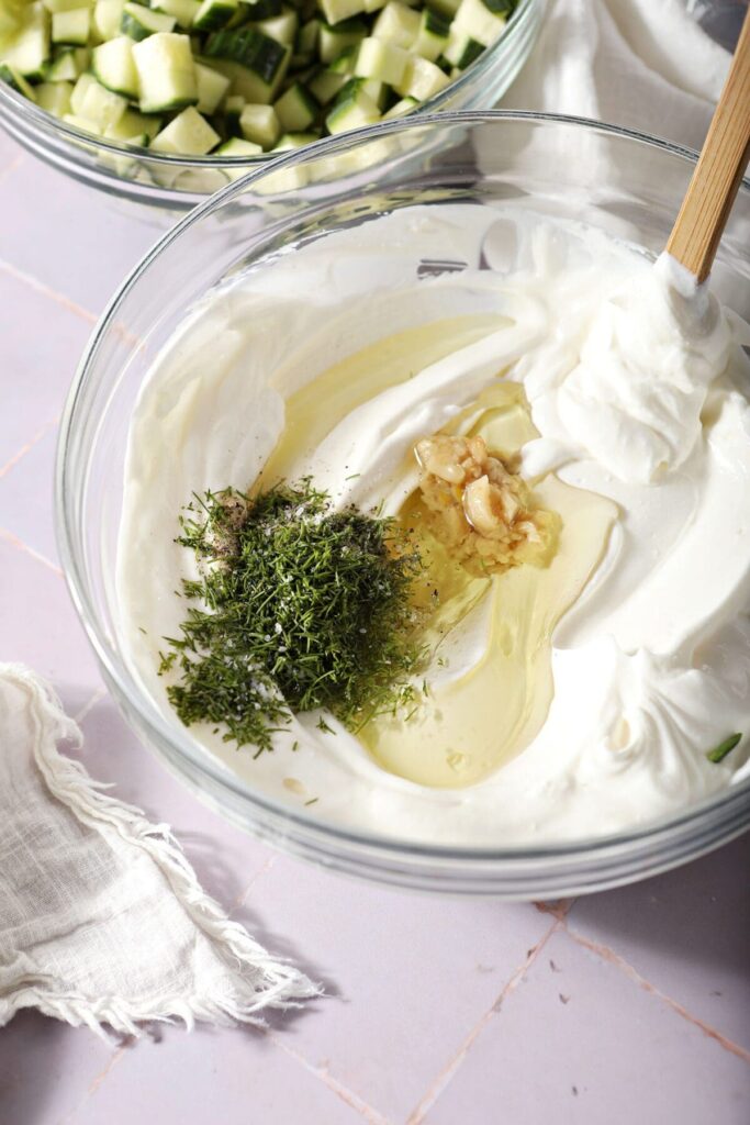 Chopped dill, salt, pepper, onion and olive oil sit on top of a bowl of sour cream and greek yogurt before stirring