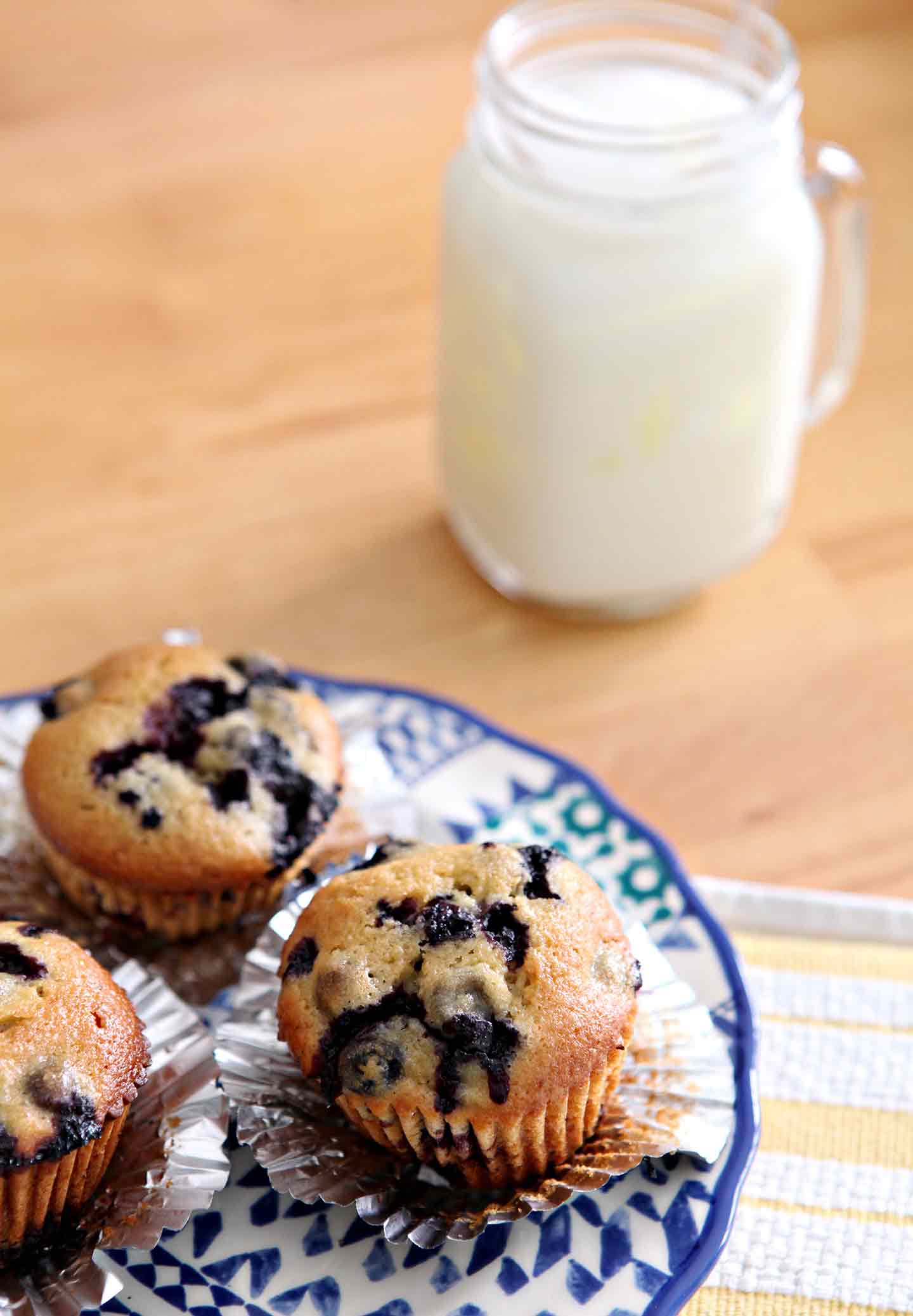 Three Vegan Blueberry Muffins, set on a blue patterned plate on a golden tabletop with a glass of nut milk before eating.