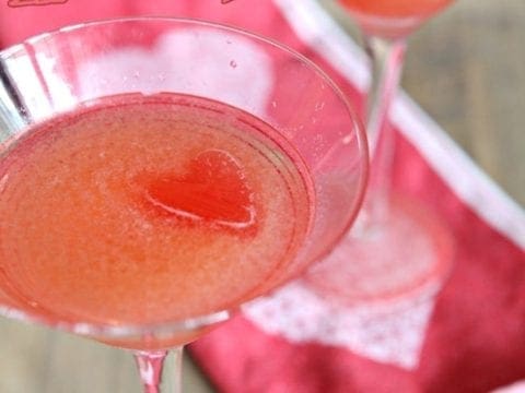 Two pink Cosmopolitan cocktails in red striped martini glasses garnished with heart ice cubes sit on a wooden surface