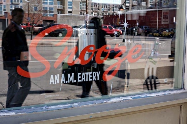 The exterior window of Snooze in Denver, showing the logo and the streetscape on the other side of it