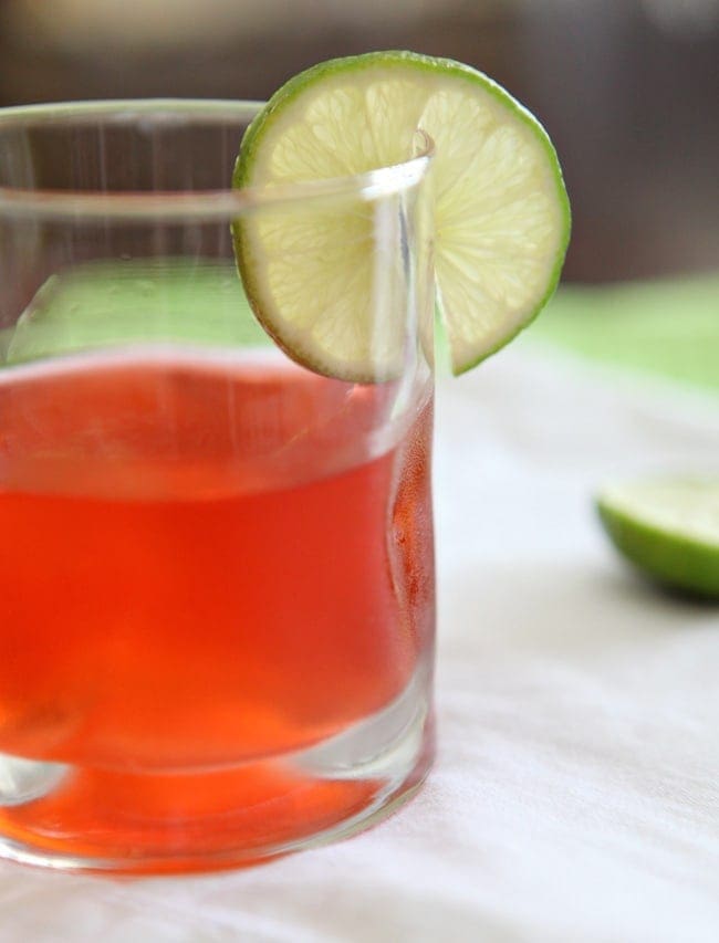 Close up of the side of a glass holding bright red liquid garnished with a lime round
