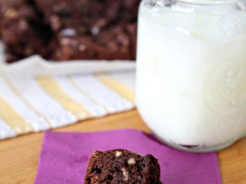A single brownie with nuts sits on a purple napkin next to a big glass of milk with more brownies in the background
