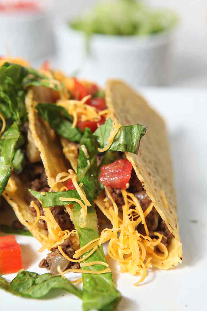 Close up of two hard shell Ground Beef Tacos, topped with lettuce, tomatoes and cheese, on a platter