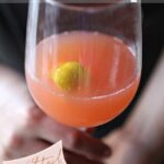 A woman holds a wine glass of a pink liquid with a lemon peel in it