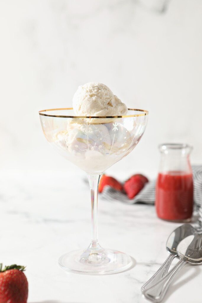 A large scoop of ice cream in a coup glass on a marble countertop next to strawberries