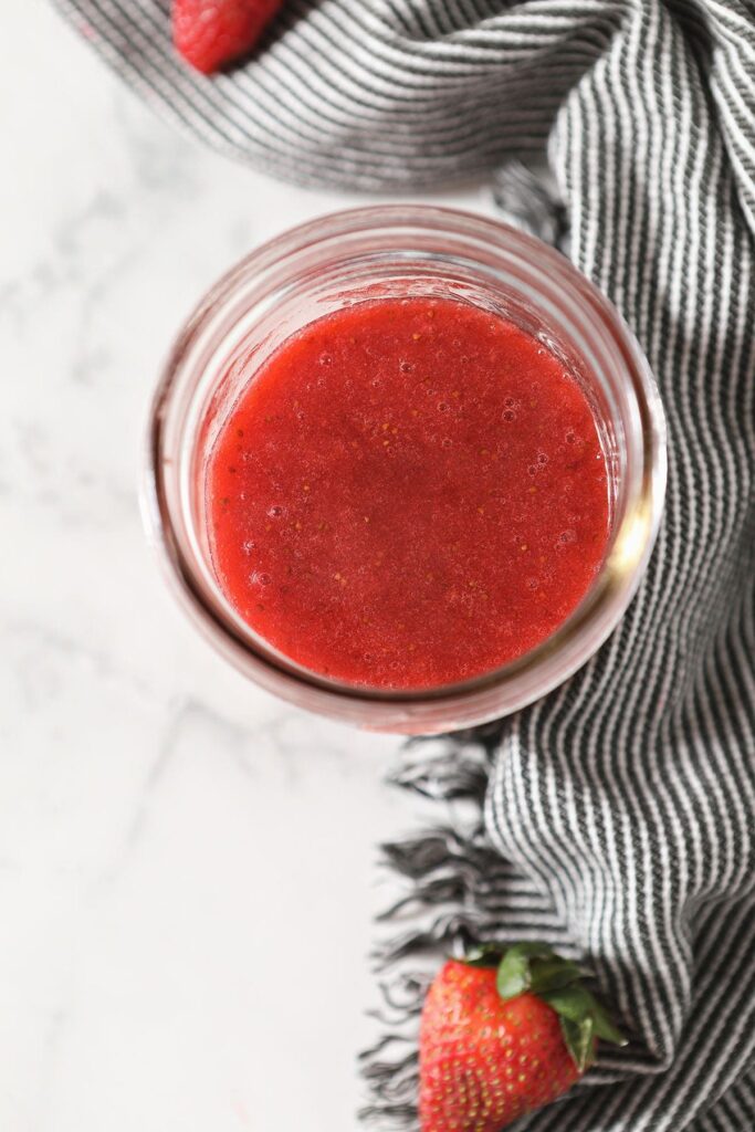 A jar of strawberry syrup on top of a marble countertop next to a dark grey striped towel