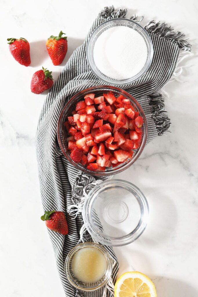 A bowl of strawberries and other ingredients in bowls sit together on a marble countertop next to a gray striped towel