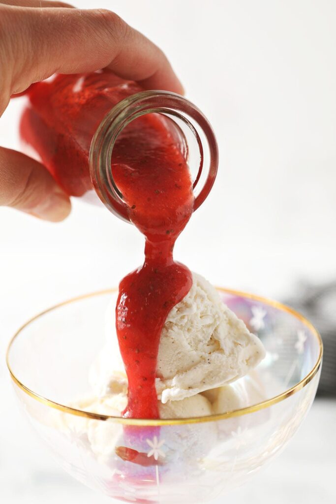 A person pours strawberry syrup on top of scoops of ice cream in a coup glass