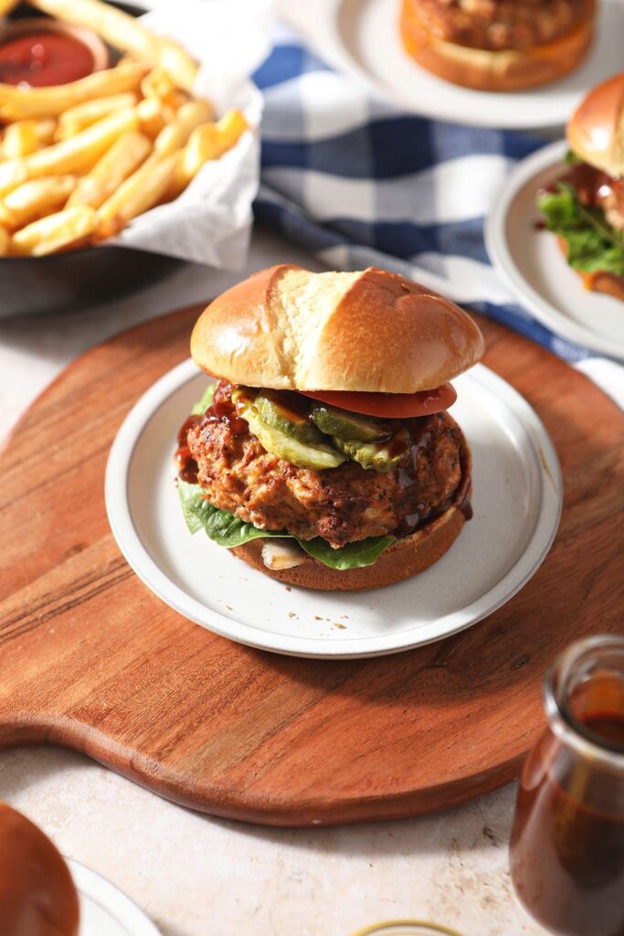 A baked BBQ turkey burger on a white plate on a wooden cutting board next to other burgers and a bowl of fries