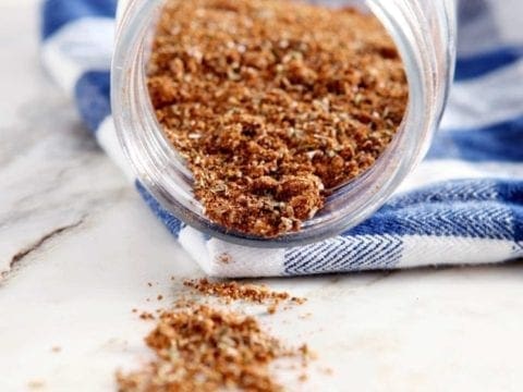 A jar of spice sitting on a blue buffalo check napkin spills onto a marble countertop