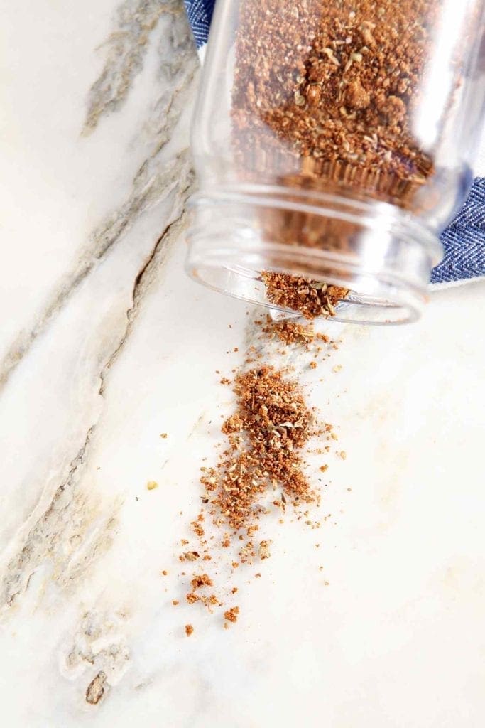 Overhead of an overturned glass jar holding a spice blend and the spice spilling out onto a marble counter