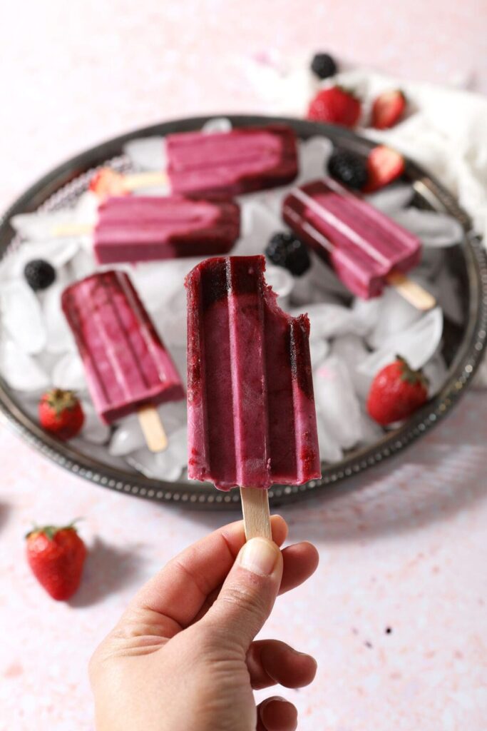 A hand holds a bitten-into ombre berry popsicle in front of a tray of more pops