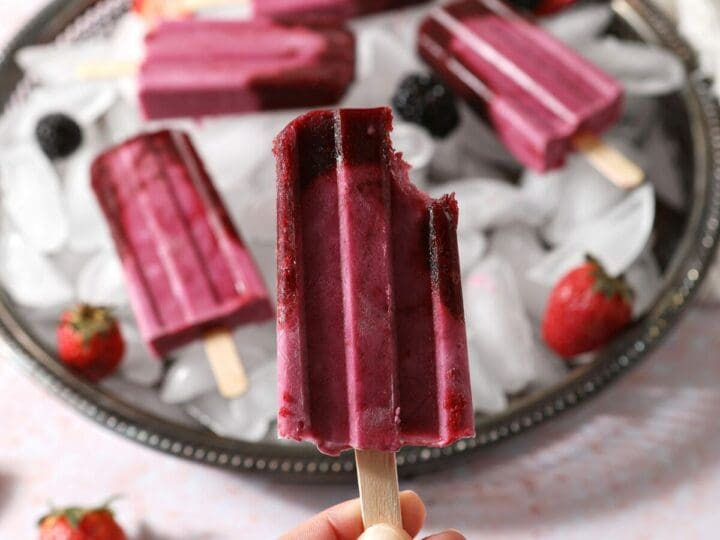 A hand holds a bitten-into ombre berry popsicle in front of a tray of more pops