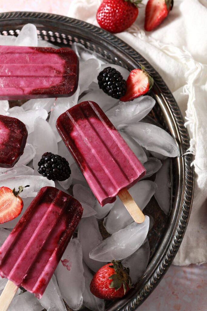 A tray of pink popsicles with berries on top of ice in a silver tray