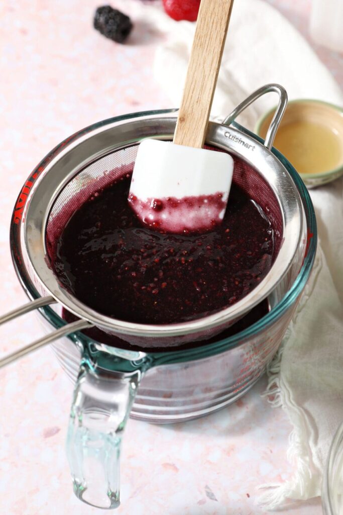 Blackberry and strawberry puree is pushed through a fine mesh strainer over a liquid measuring cup