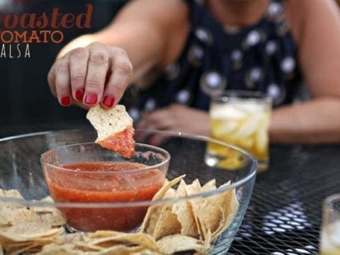 A woman with red nails dips a tortilla chip into a bowl of salsa