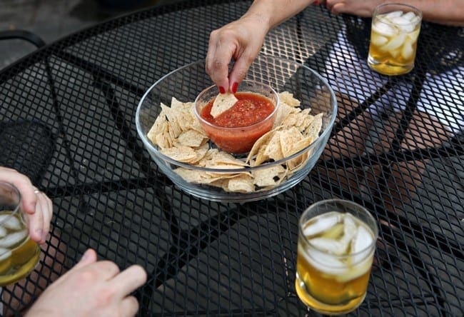 A woman with red nails dips a tortilla chip into a bowl of salsa while surrounded by three glasses of a golden liquid