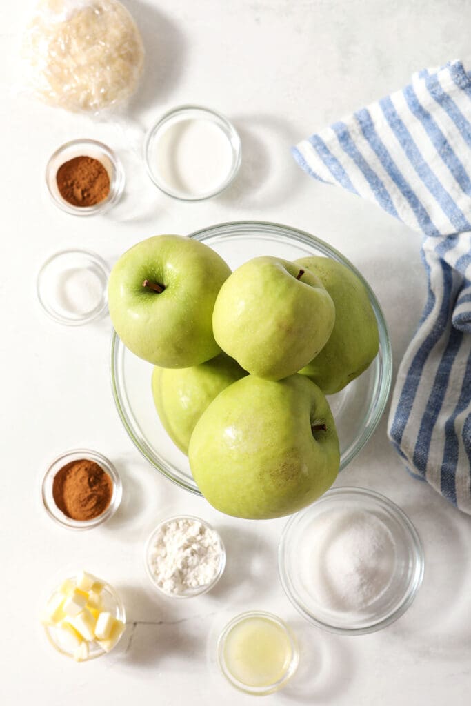 Ingredients to make apple pie with Granny Smith apples in bowls on a white surface with a blue and white striped linen