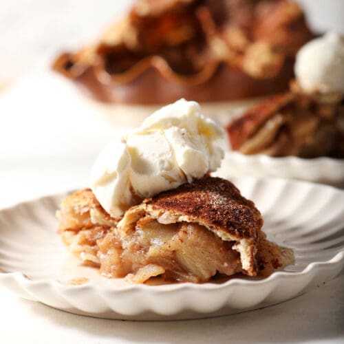Side angle of a slice of apple pie topped with vanilla ice cream in front of the rest of the pie on a white surface