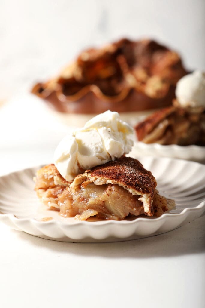 Side angle of a slice of apple pie topped with vanilla ice cream in front of the rest of the pie on a white surface