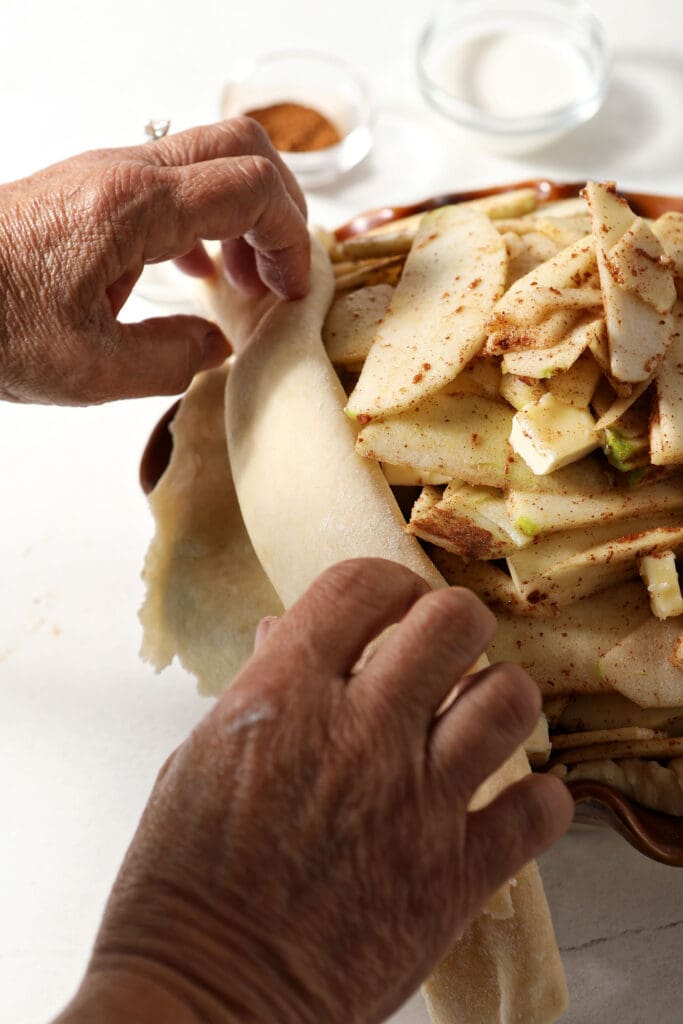 A hand rolls pie dough on top of apples in a pie dish before baking