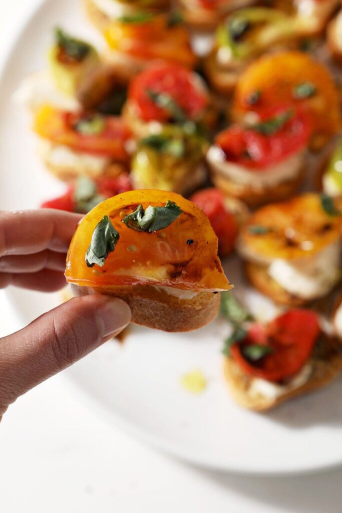 A hand holds a Caprese Bruschetta in hand above a white platter