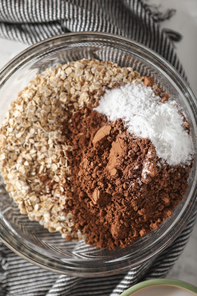 Rolled oats, cocoa powder, salt and baking powder in a bowl on top of a gray and white striped towel