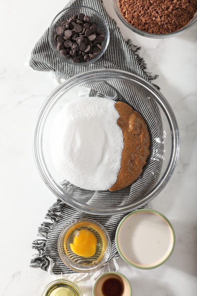 Granulated sugar and almond butter in a large glass bowl on a gray and white striped towel surrounded by other cookie ingredients