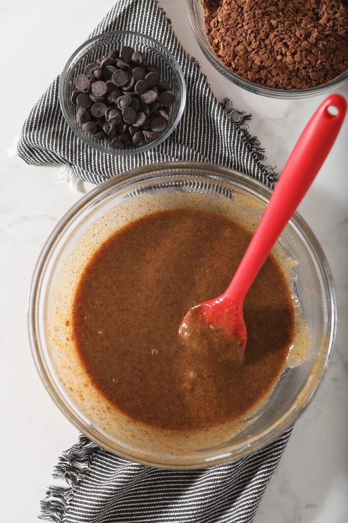 The cookie wet ingredients are in a clear glass bowl with a red spatula next to a bowl of chocolate chips and a bowl of oats coated in cocoa powder