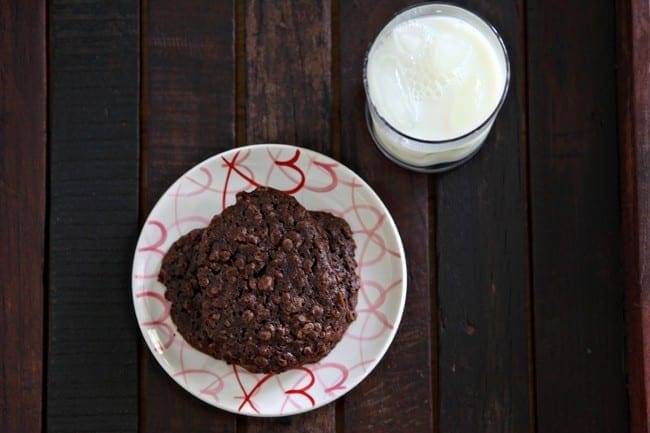 A stack of Gluten Free Chocolate Cookies on a white plate with pink and red hearts on it, sitting on a wooden surface with a glass of milk