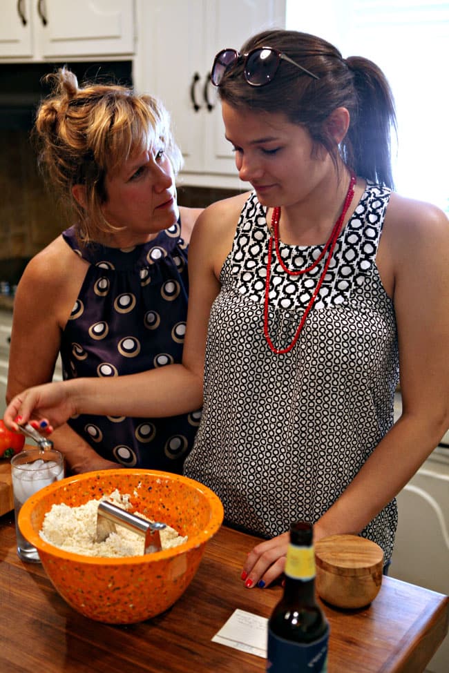 A blond woman and a brunette woman stand together in front of an orange bowl holding pie crust ingredients as they make a pie