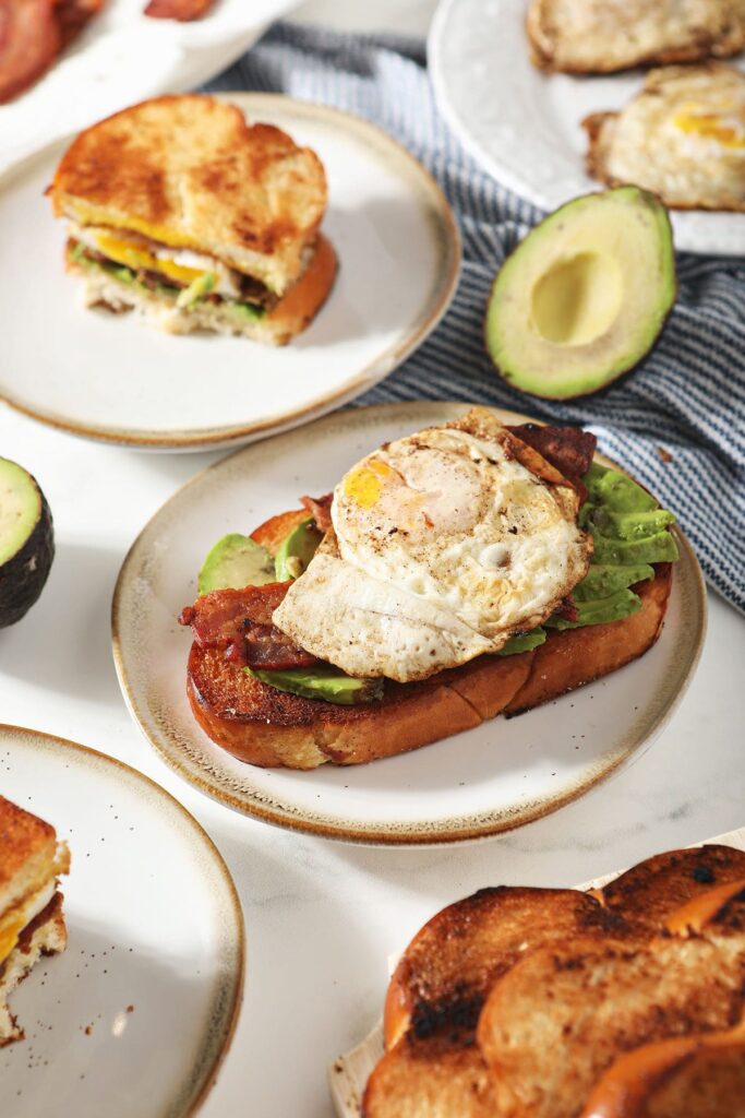 Open-faced breakfast sandwiches on small plates with egg, bacon and avocado