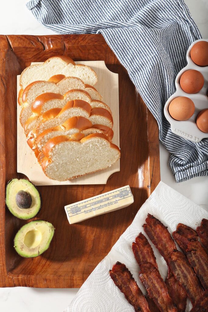 Sliced bread, avocado, eggs and bacon to make breakfast sandwiches on a cutting board