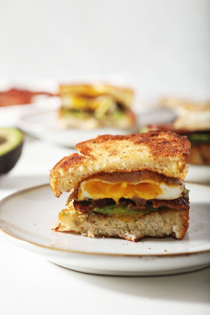 A halved Bacon Avocado Egg Sandwich on a white plate with a runny yolk