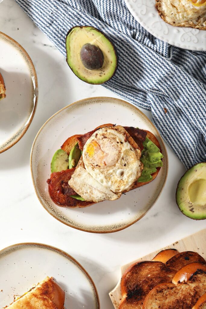 An open-face Bacon Avocado Egg Sandwich on a white plate surrounded by other sandwiches and halved avocados