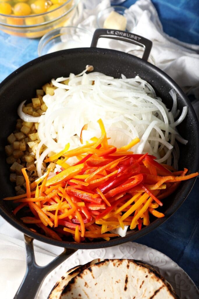 Sliced sweet peppers and white onion in a skillet on top of potatoes