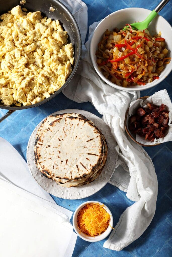 Scrambled eggs, cooked potatoes with onions and peppers, warmed tortillas, cooked bacon and shredded cheese in bowls on a blue surface