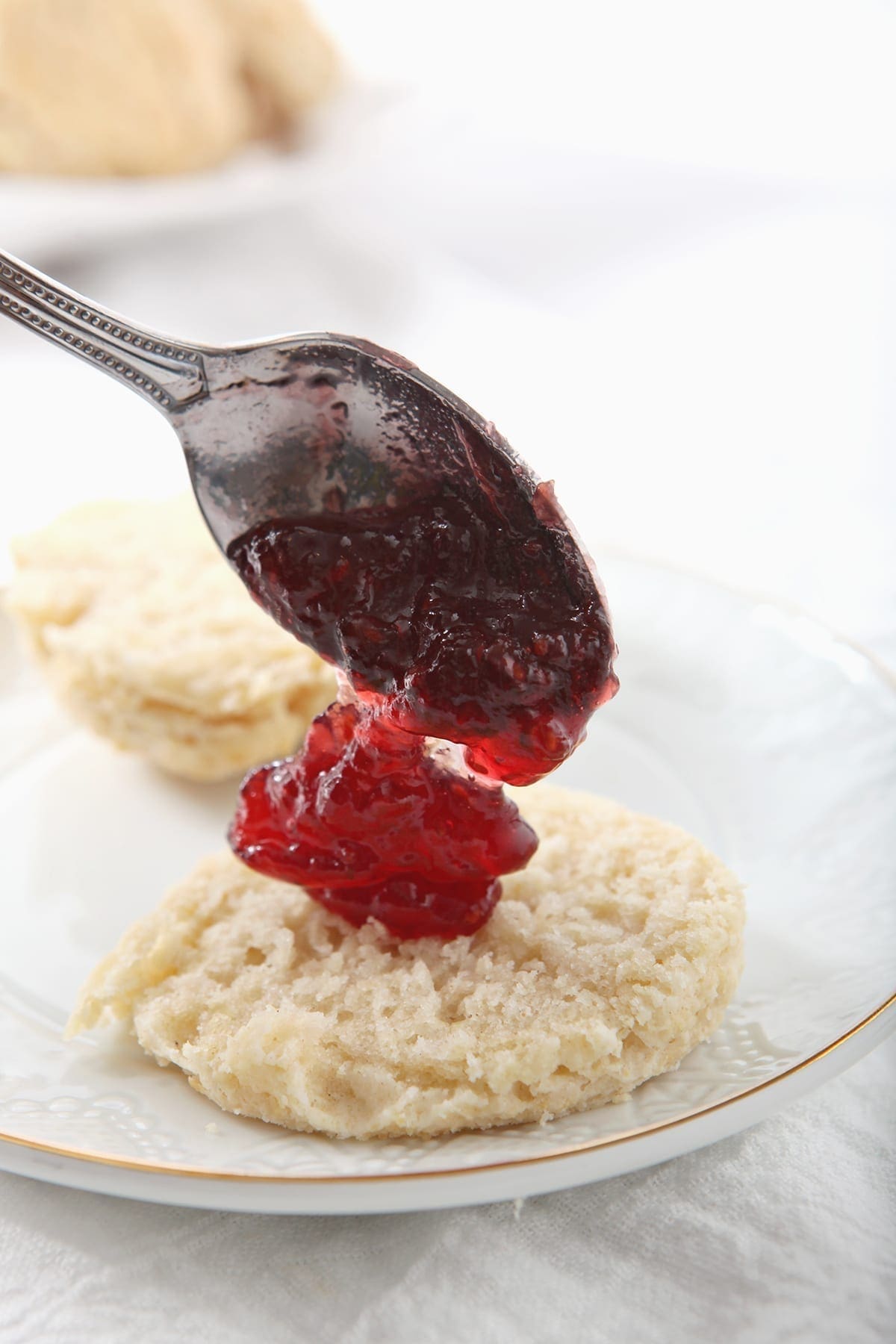 A spoon dollops raspberry jam into the middle of a vegan biscuit