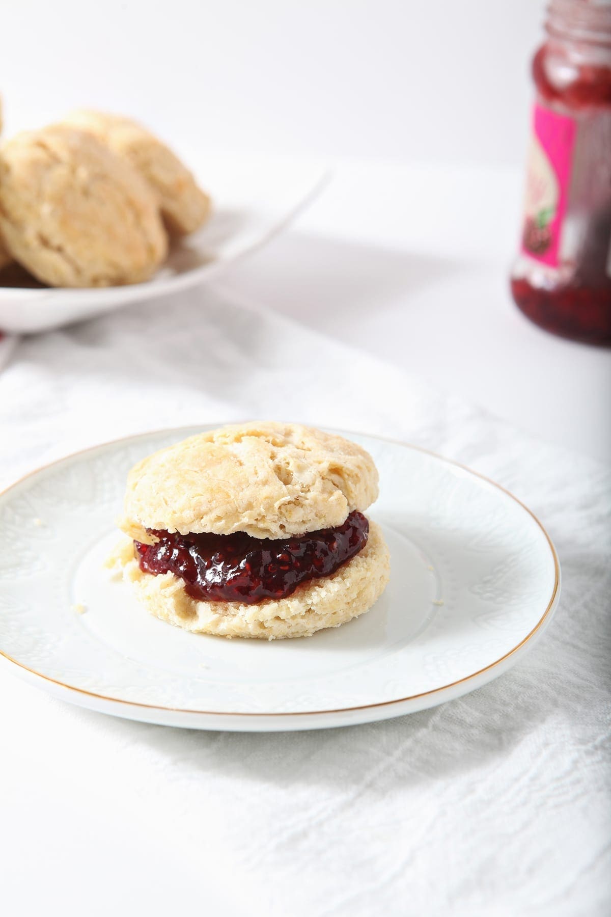 A biscuit with raspberry jam on a white plate with other biscuits and jam in the background