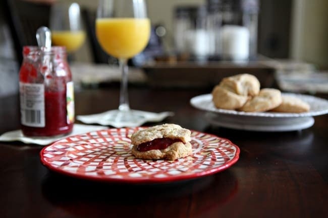 A side view of a biscuit with jam on a red plate on a dark wooden tabletop with mimosas, a jar of jam and other biscuits