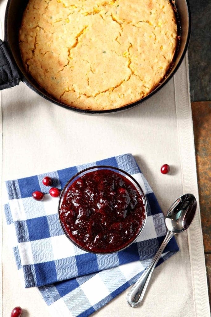 An overhead of a glass bowl of Spiced Cranberry Sauce on a buffalo check towel with a spoon next to cornbread