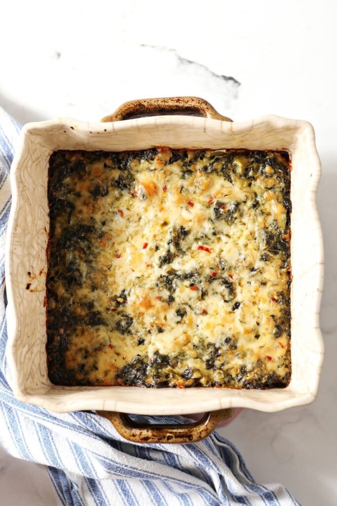 A casserole dish holding Baked Creamy Kale on marble with a blue striped towel