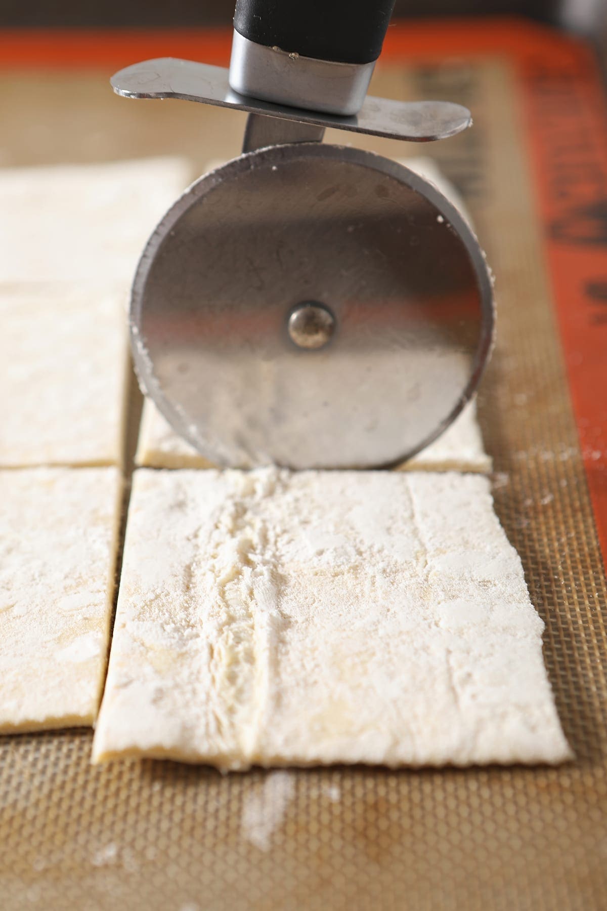 A pizza cutter slices a sheet of puff pastry into squares on a Silpat-lined baking sheet