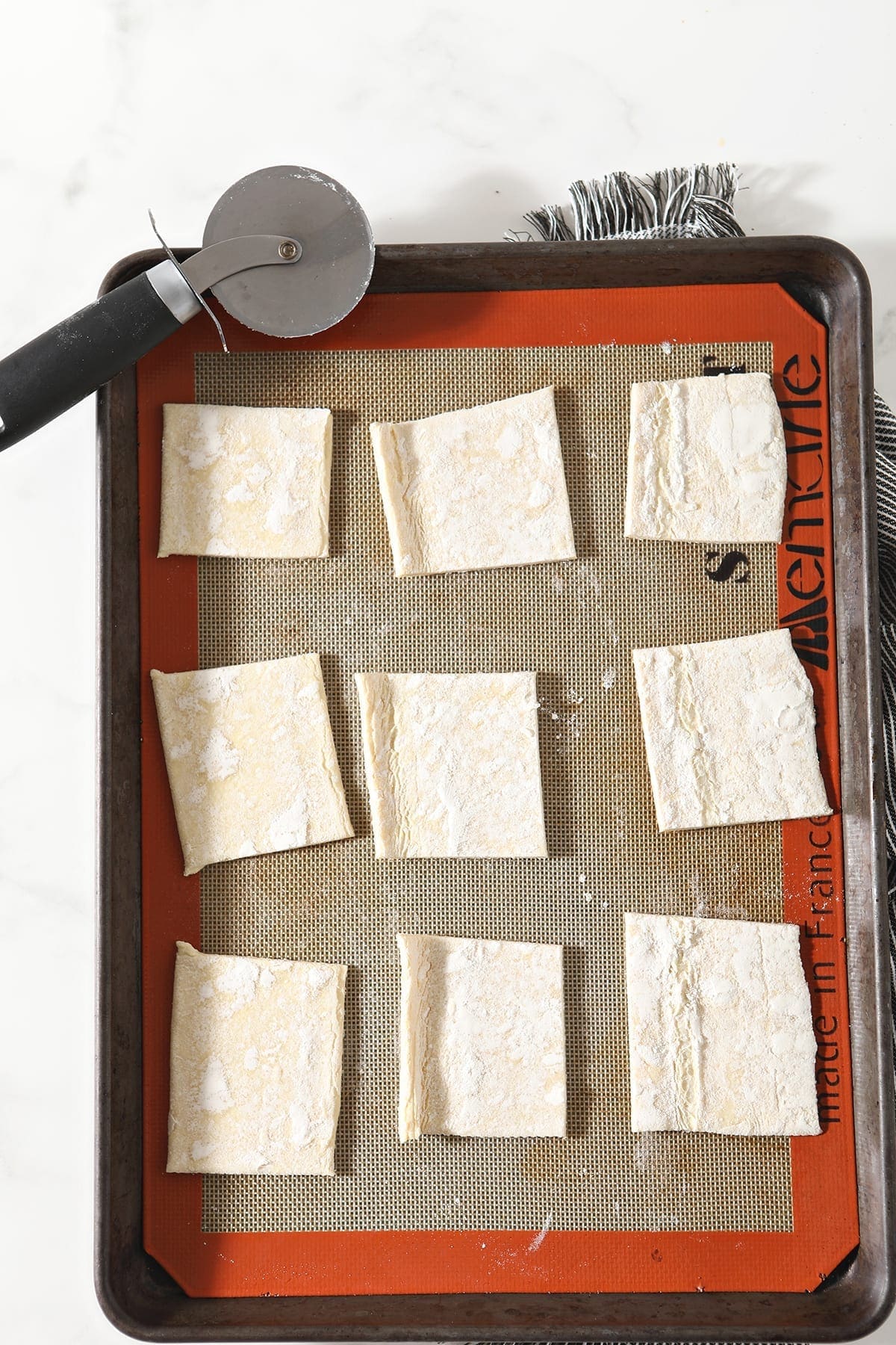 Squares of puff pastry on a Silpat-lined baking sheet with a pizza cutter