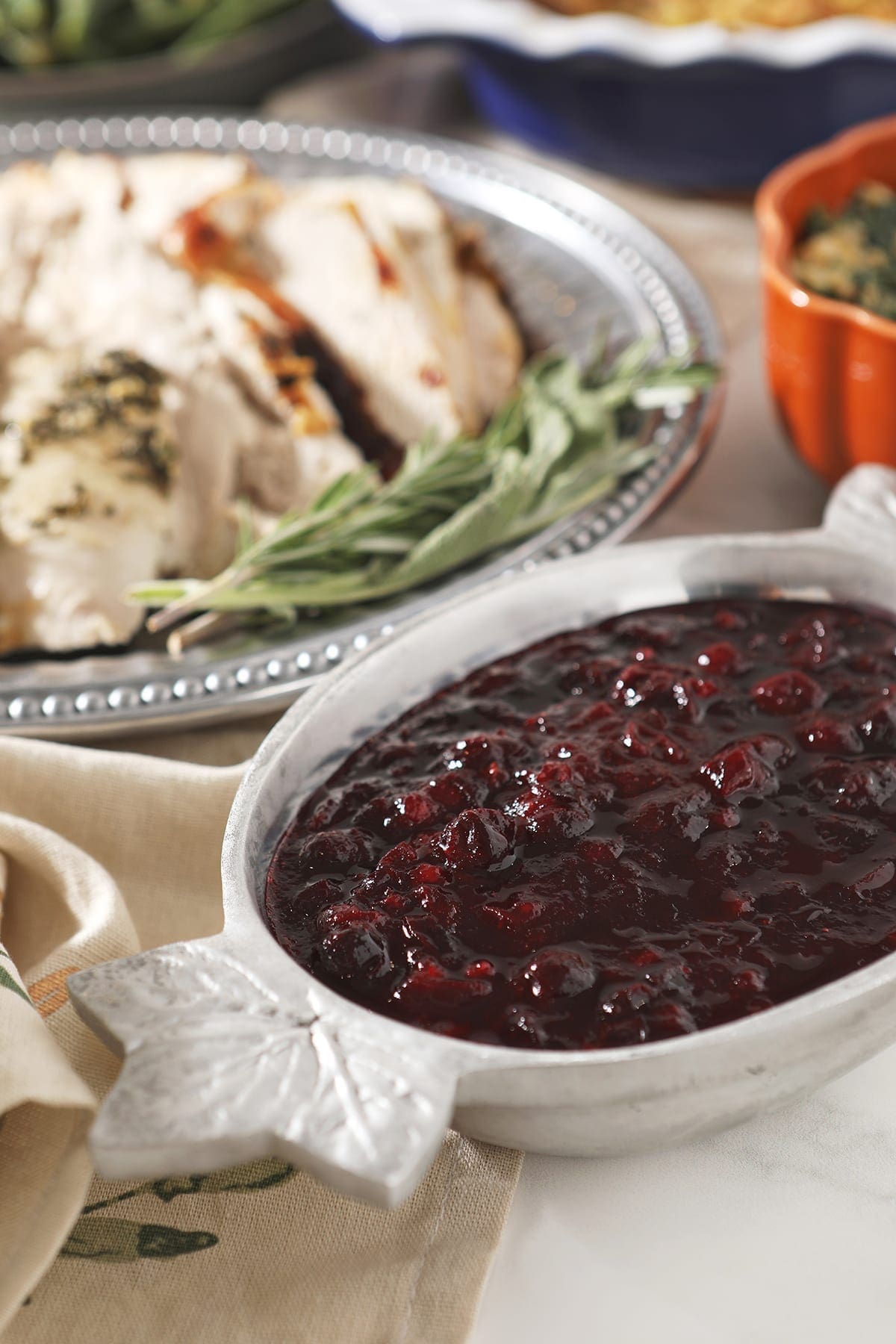 Cranberry sauce in a silver bowl with leafs on the ends next to a silver platter of turkey