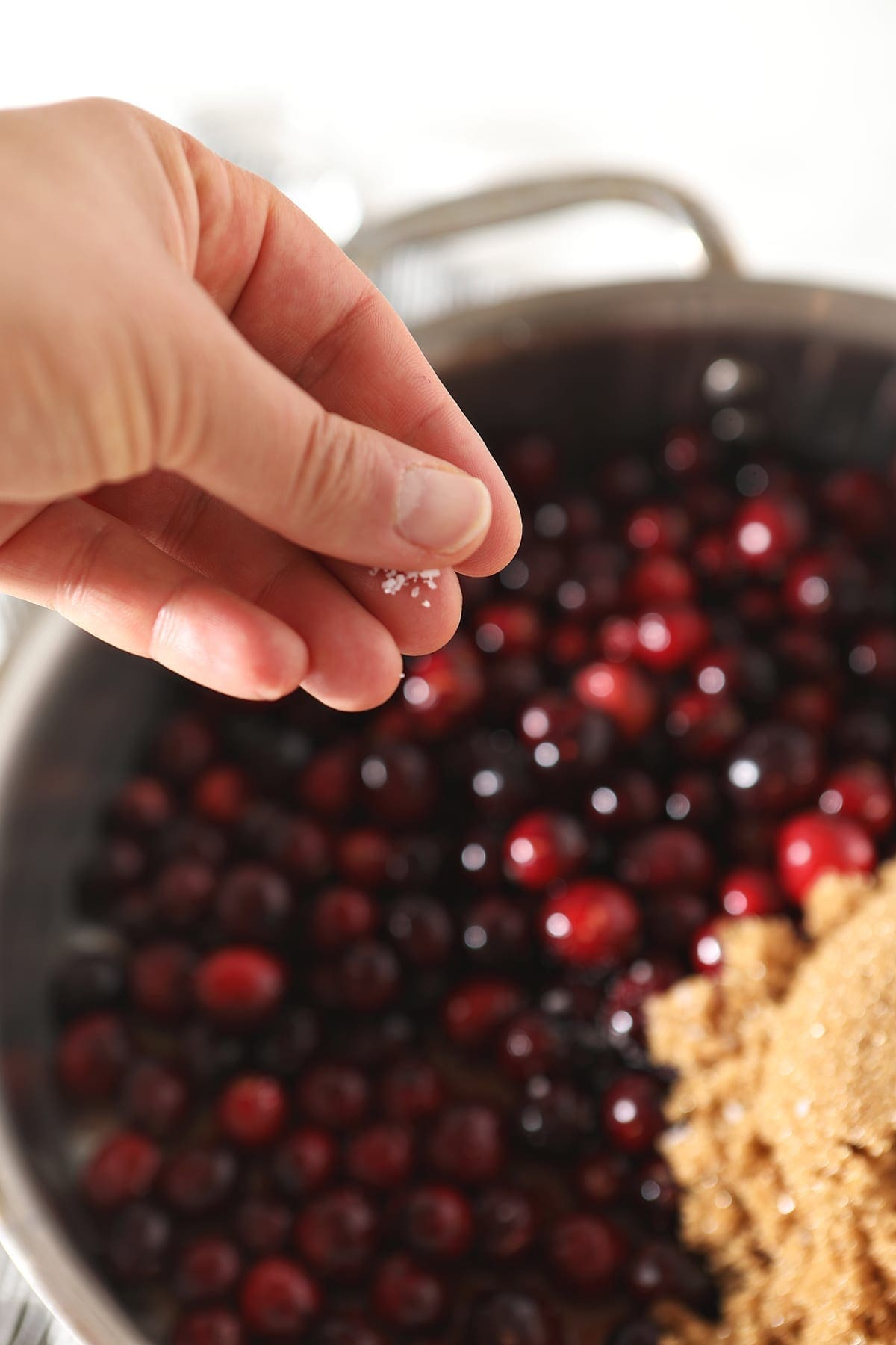 A hand sprinkles salt on top of cranberries and brown sugar in a skillet