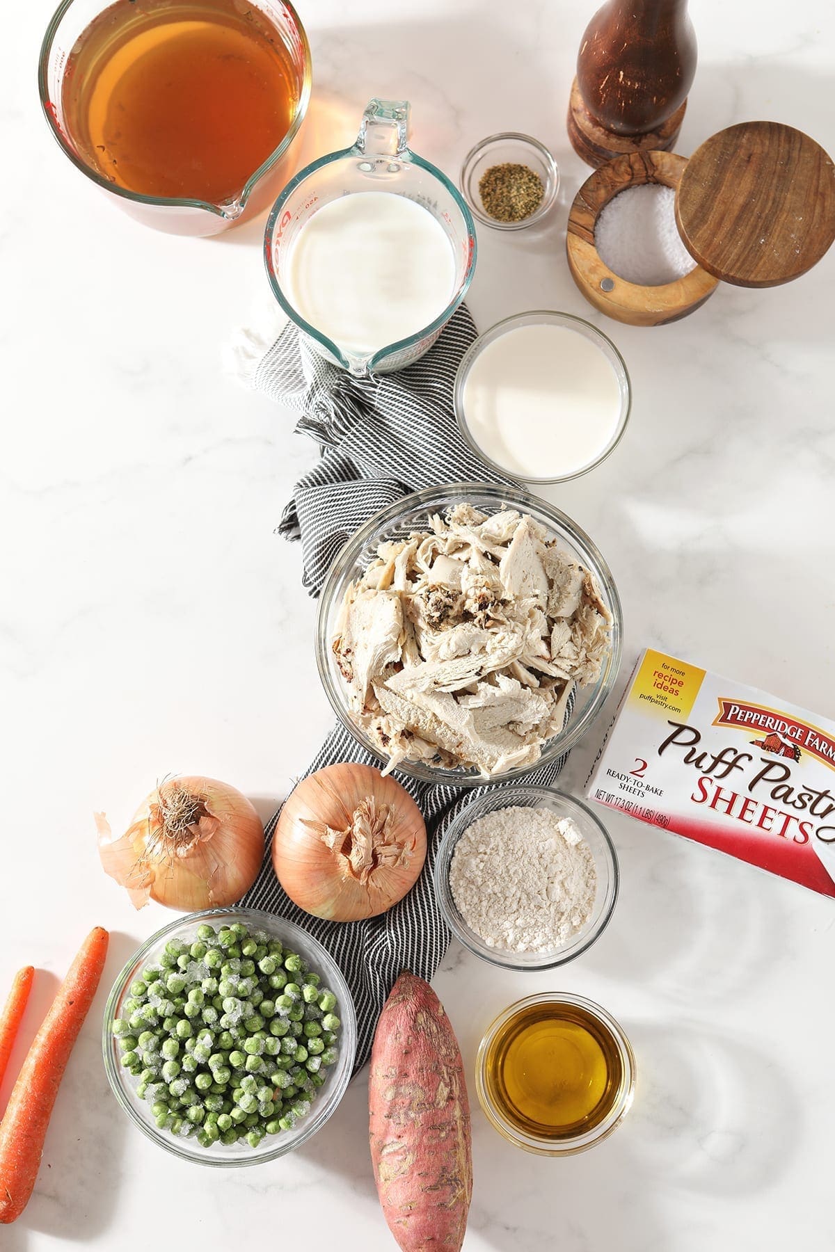 Shredded leftover turkey breast, vegetables, stock and milk, among other ingredients, sit on a gray striped towel on a marble countertop in bowls