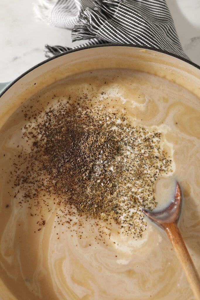 Spices are shown on top of the pot pie liquid in a Dutch oven with a wooden spoon before stirring