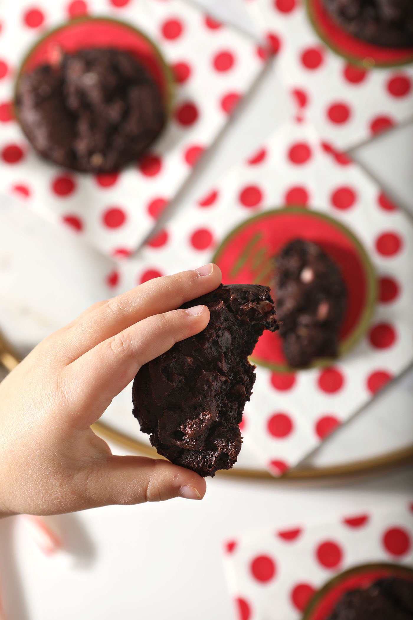 A child holds a halved Chocolate Peppermint Chocolate Chip Cookie above more cookies on red and white polka dot napkins